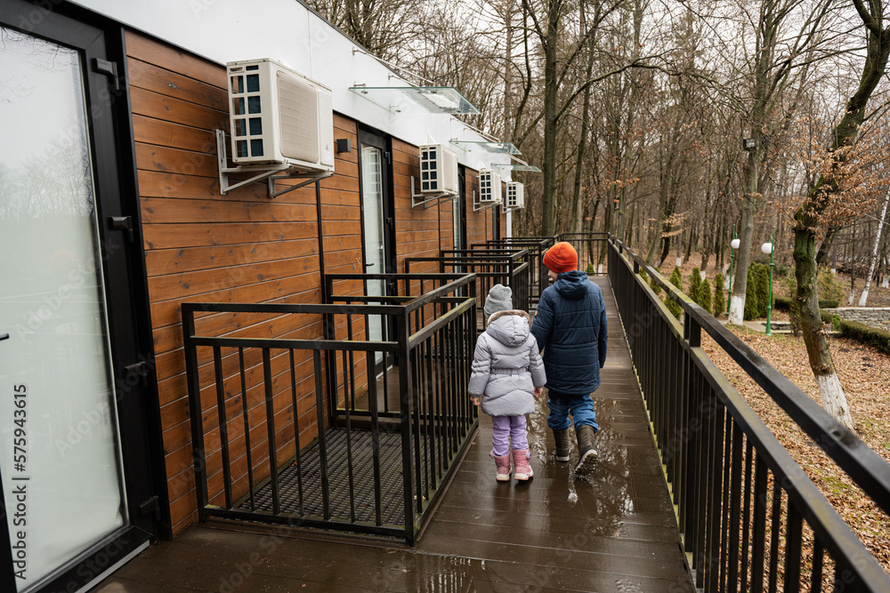 Back of brother with sister walking on terrace of one-storey modular houses in spring rainy forest.