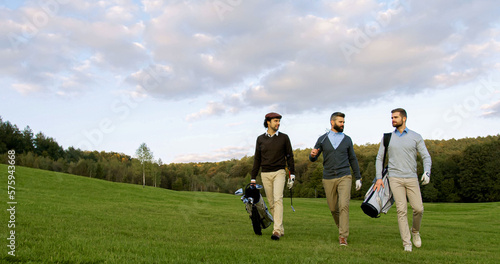 Three handsome men strolling with their clubs and golf bags on the golf field to start a game and talking. Outdoor