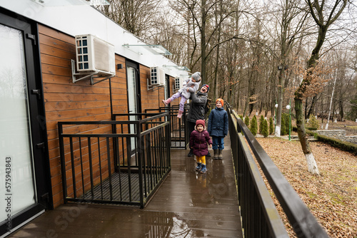 Father with three kids on terrace of one-storey modular houses in spring rainy forest.