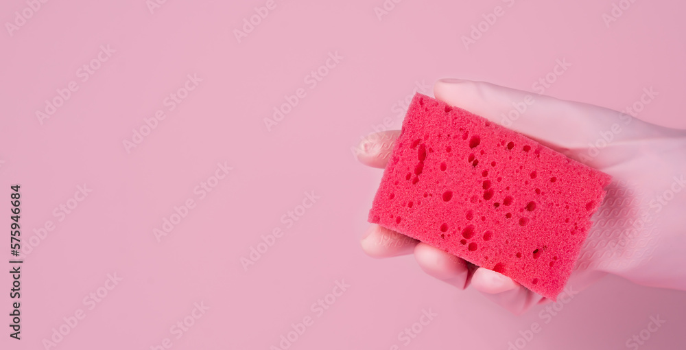A woman's hand in a rubber glove holds a cleaning sponge on a pink background. Washing and cleaning concept. Close-up. Copy space. Banner. Selective focus. A woman's hand in a rubber glove holds a cleaning sponge on a pink background. Washing and cleaning concept. Close-up. Copy space. Banner. Selective focus.