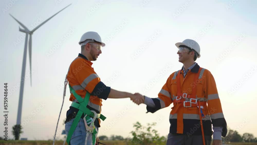 Two caucasian engineers man in hardhat and goggles talk and say goodbye with shaking hands before leaving work at agricultural field with wind turbines before sunset, wind turbine generate electricity Two caucasian engineers man in hardhat and goggles talk and say goodbye with shaking hands before leaving work at agricultural field with wind turbines before sunset, wind turbine generate electricity