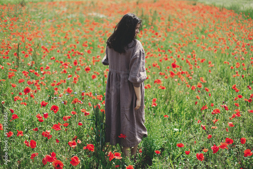 Stylish woman in rustic dress walking in red poppy field. Cottagecore aesthetics. Young female in linen dress relaxing among wildflowers meadow in sunny summer countryside, rural slow life