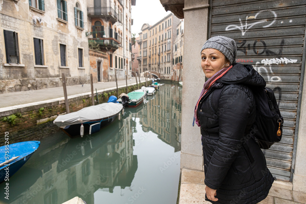 A young backpacker woman in winter clothes visiting Venice's canals. Boats on water and graffiti on the wall