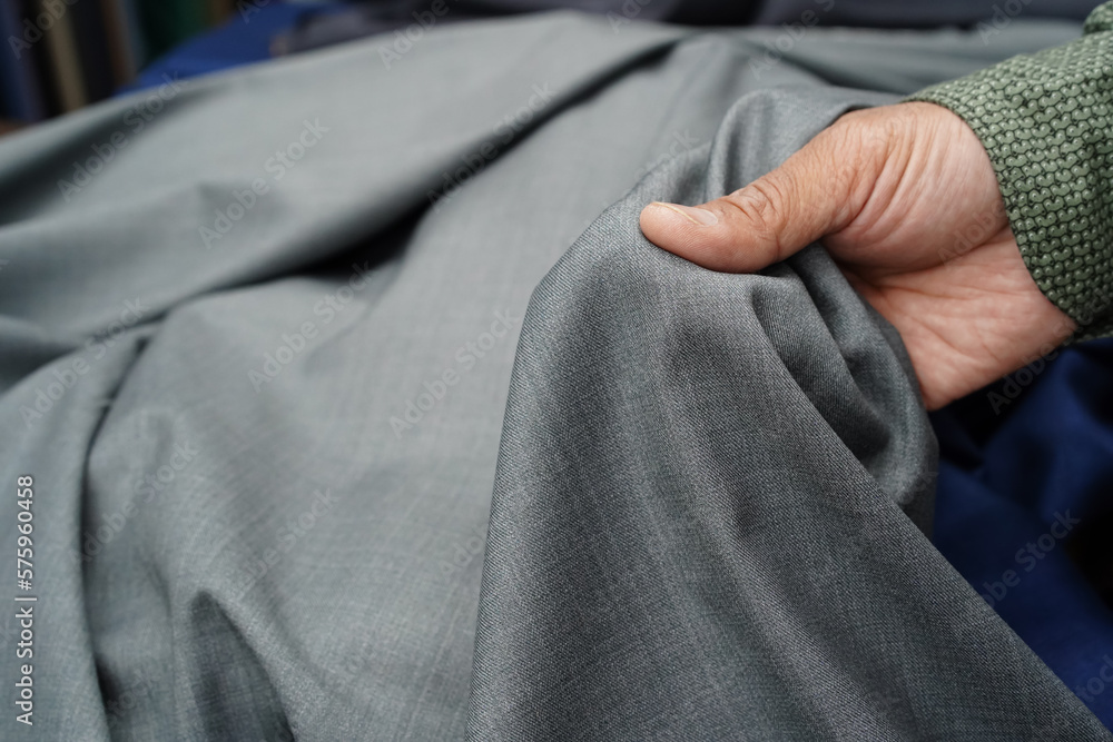 A man's hand holding a piece of quality silver grey fabric. Choosing a textile material in a tactile and visual way to tailor a garment, trouser, jacket. Close-up, selective focus, blurred background.