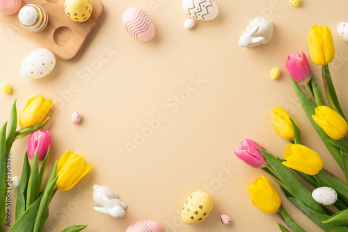 Easter decor concept. Top view photo of colorful easter eggs ceramic bunnies yellow and pink tulips and wooden egg holder on isolated pastel beige background with copyspace in the middle