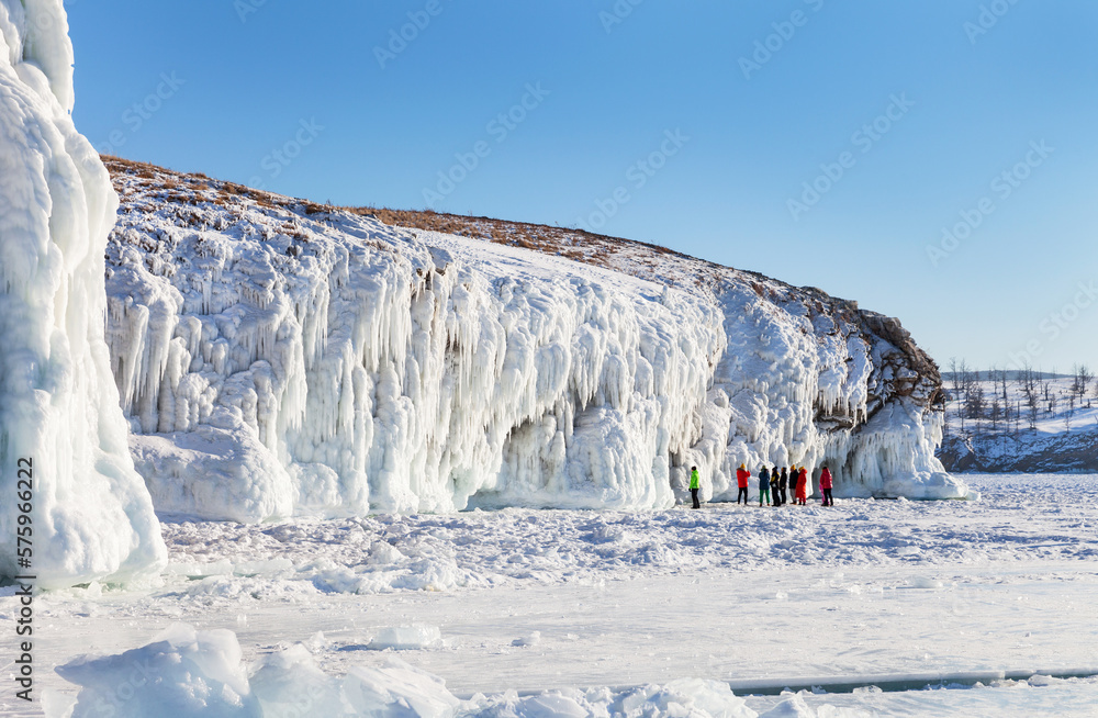 Fototapeta premium Frozen Baikal Lake. In winter, coastal cliffs of Olkhon Island are covered with white crust of splashing ice with long icicles and bizarre ice grottoes. Group of tourists inspecting scenic icy rocks