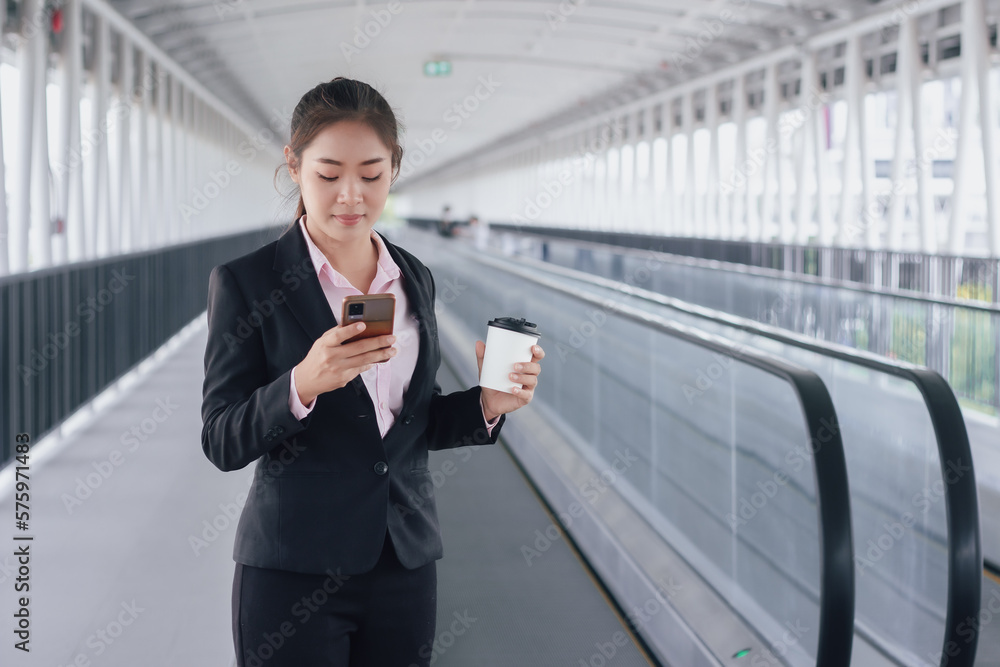 young Asian businesswoman in black suit using phone and holding cup of coffee and standing on elevated walkway station with copy space. young Asian businesswoman in black suit using phone and holding cup of coffee and standing on elevated walkway station with copy space.