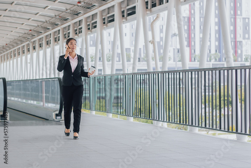 young Asian businesswoman in black suit using phone and holding cup of coffee and standing on elevated walkway station with copy space.