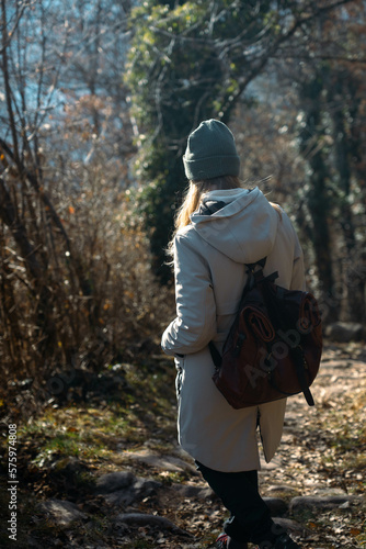 Girl with hat and leather backpack walks on a mountain road. Female hiker in winter walks in nature. Isolation, solitude, peace and silence.