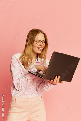Happy young pretty blonde girl, student wearing modern office style clothes using laptop over pink background. Concept of youth, student college life, business and education.