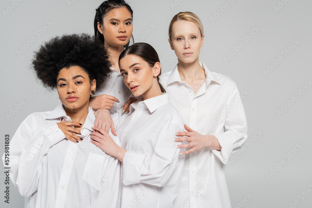 sensual multicultural women in white shirts looking at camera while standing isolated on grey. sensual multicultural women in white shirts looking at camera while standing isolated on grey.