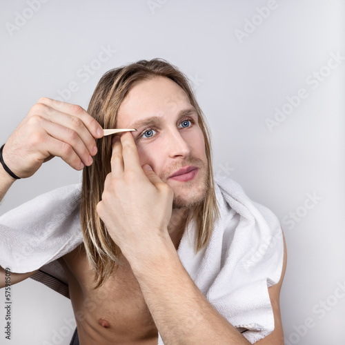 A smiling topless young long-haired blond with blue eyes is doing his own eyebrow correction with tweezers,he has a white towel on his shoulders.Cosmetology for men