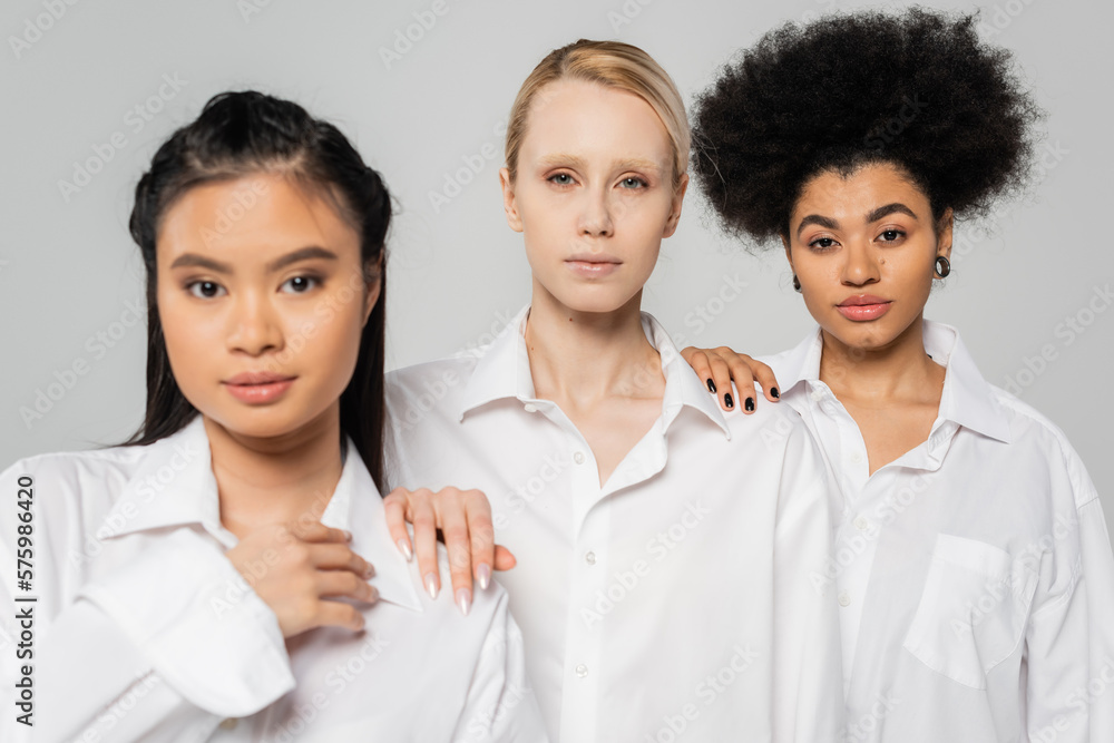 young blonde model looking at camera near asian and african american women in white shirts isolated on grey. young blonde model looking at camera near asian and african american women in white shirts isolated on grey.
