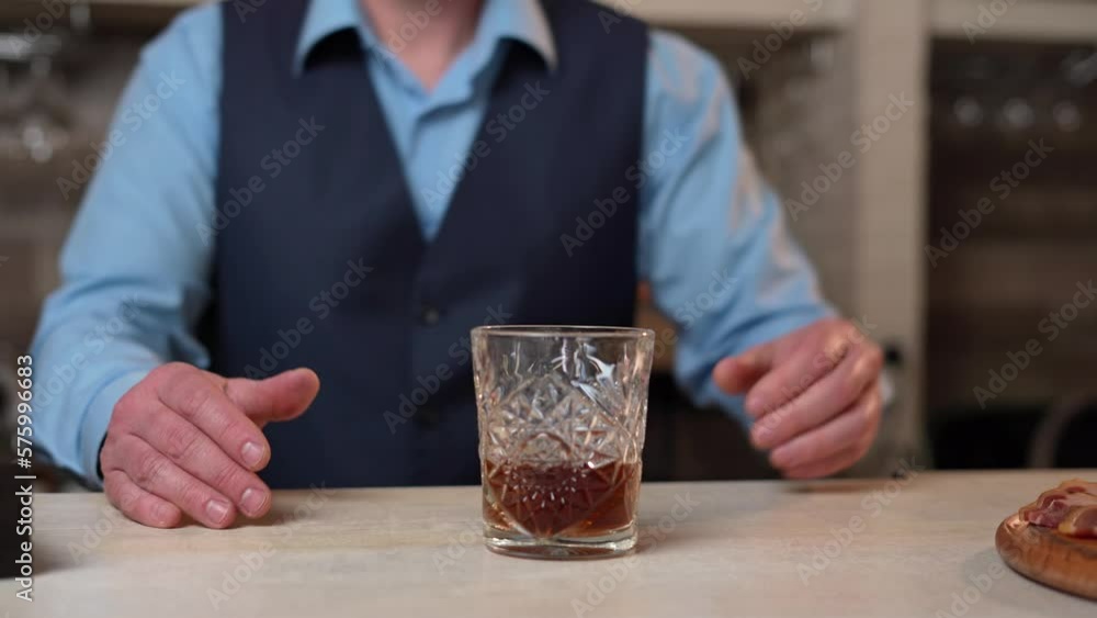 A Close-up of the hands of the Bartender, that Removes the Whisky Bottle from the Bar counter. A Bartender in a blue shirt and black vest stands behind the Bar counter Preparing a Cocktail A Close-up of the hands of the Bartender, that Removes the Whisky Bottle from the Bar counter. A Bartender in a blue shirt and black vest stands behind the Bar counter Preparing a Cocktail