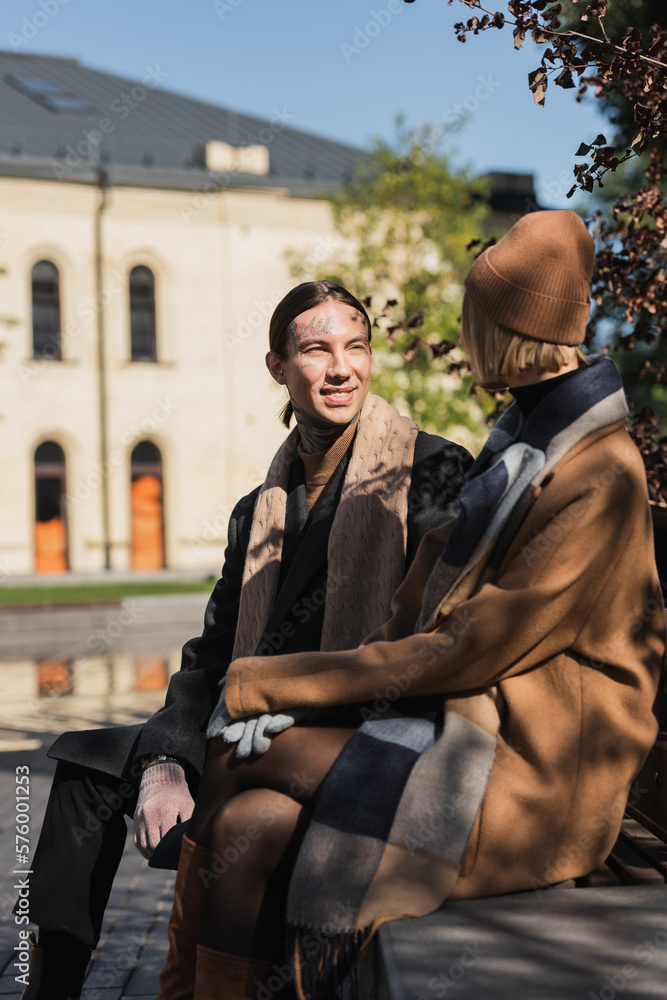 Naklejka premium happy tattooed man looking at blonde woman in hat while sitting on bench.