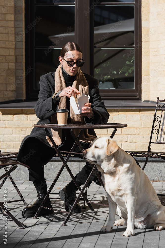 full length of tattooed man in sunglasses holding chopsticks while eating asian food near dog in outdoor cafe.