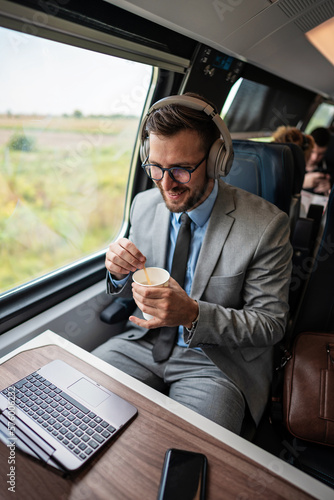 Handsome businessman is having a good time while traveling by high-speed train. He is using laptop computer and wireless headphones for online communication, gaming and entertainment. © Dusko
