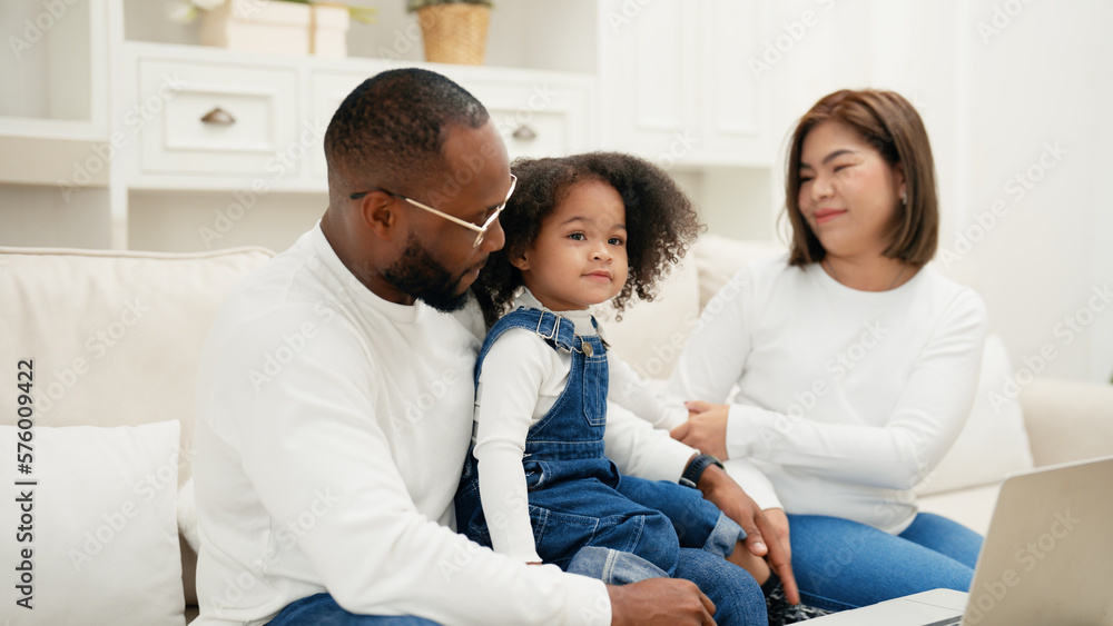 Multiracial newbie parent is sitting on a sofa with their daughter and giving love to her while watch online entertainment contents on a laptop. African adult man is kissing his daughter to show love.