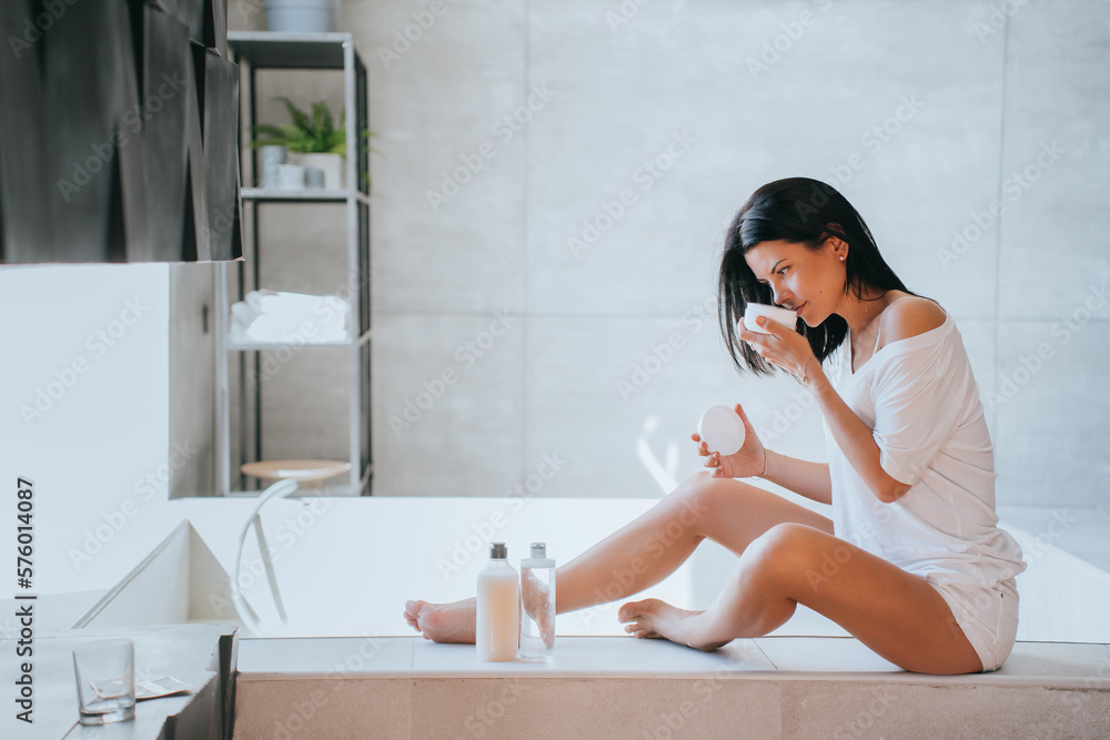 Beautiful brunette hispanic young adult woman sitting at bathroom smells new skincare cream, spa procedure. Caucasian housewife trying different lotions for legs, moisturiser. Health care. Beautiful brunette hispanic young adult woman sitting at bathroom smells new skincare cream, spa procedure. Caucasian housewife trying different lotions for legs, moisturiser. Health care.