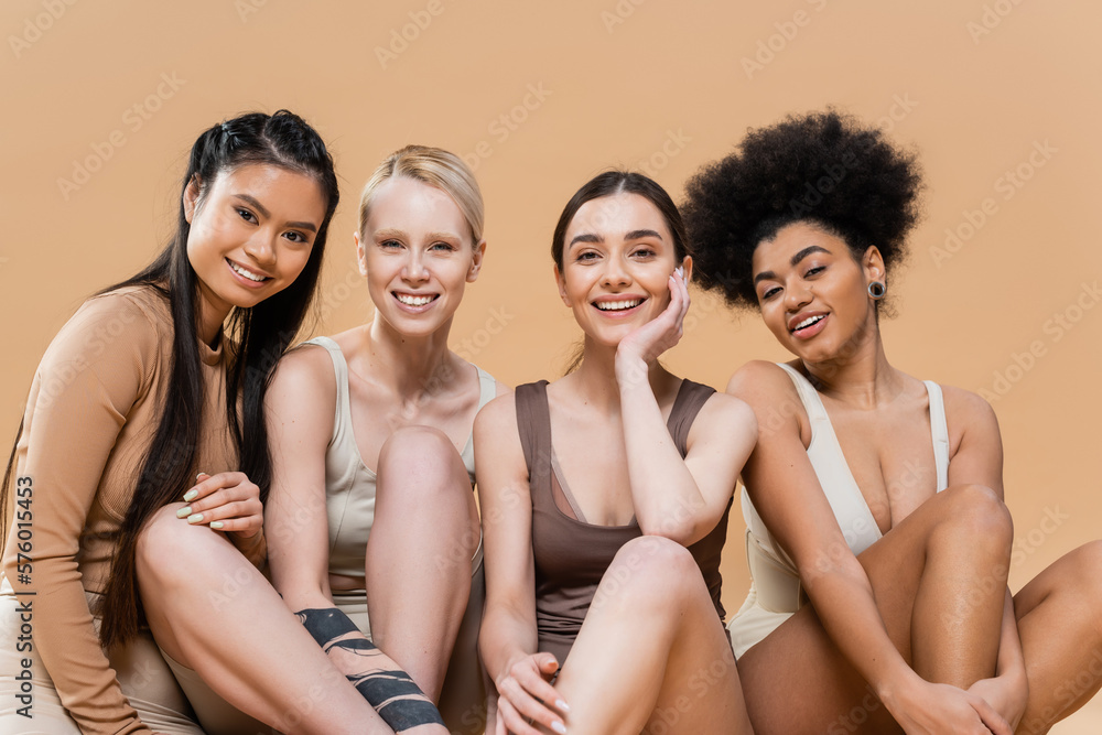 pleased multicultural models in underwear sitting and smiling at camera isolated on beige. pleased multicultural models in underwear sitting and smiling at camera isolated on beige.