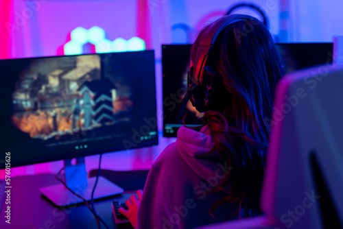 Back of a woman with confidence is going live or streaming during a game. She wears headphones and speaks to her teammates or those who are watching her on social networks or online platforms.