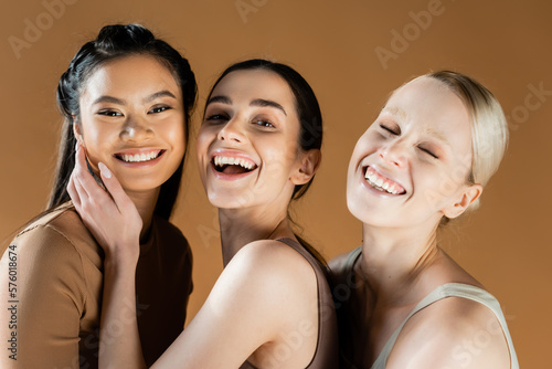 young and happy multicultural women in underwear smiling at camera isolated on beige.
