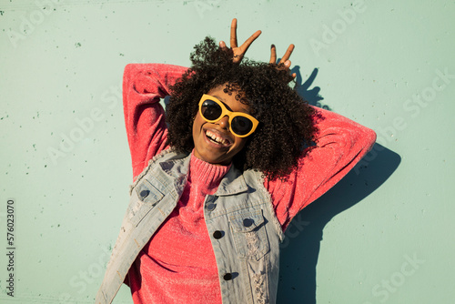 Cheerful young woman wearing sunglasses showing peace gesture in front of wall photo
