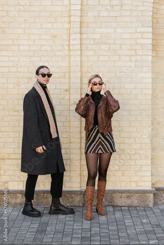 full length of stylish woman adjusting sunglasses and standing near brick wall with tattooed man.