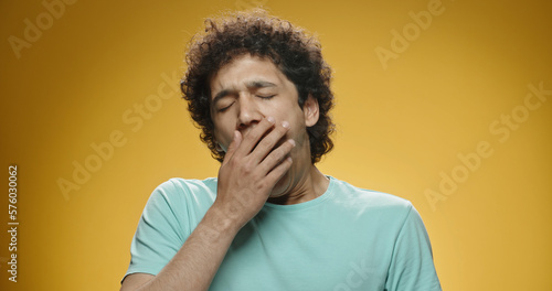 Young asian man with curly hair in casual clothes beind tired or bored, yawning and covering his mouth, isolated on yellow background close up  © andreybiling