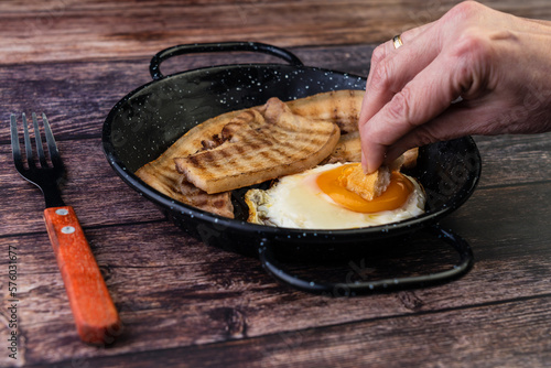 sarten de huevos con panceta encima de una mesa de madera, mujer mojando pan en la yema del huevo photo