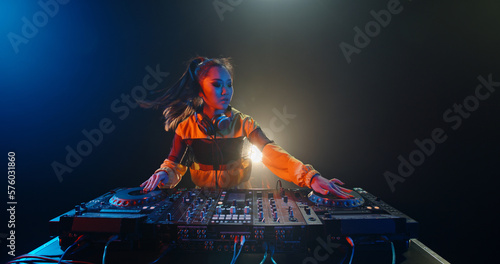 Cool female asian dj is working in a nightclub, standing at turntables, creating a dance music set - nightlife concept 