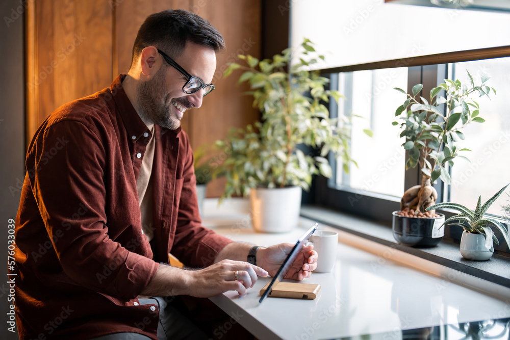 Satisfied millennial man using digital tablet sitting at table near window in his apartment. Young handsome guy using apps on tablet technology device reading, ordering online or browsing internet. Satisfied millennial man using digital tablet sitting at table near window in his apartment. Young handsome guy using apps on tablet technology device reading, ordering online or browsing internet.