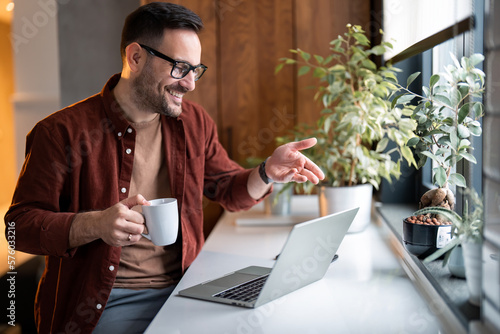 Photos Cheerful handsome confident man having remote online work hybrid meeting or distance job interview, holding cup of coffee, gesturing with hands during virtual video conference call from home