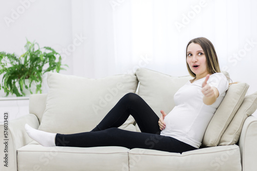 Cute pregnant woman showing thumb up sitting on the sofa