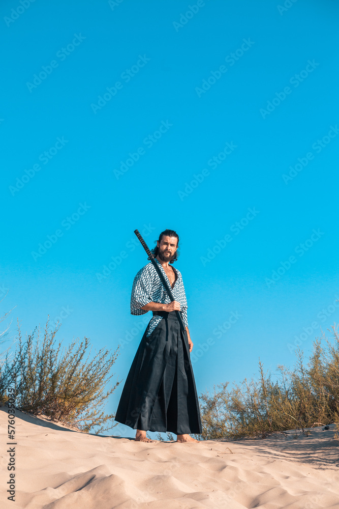 Fototapeta premium A Japanese warrior in a traditional kimano armed with a katana sword on a sandy shore next to the ocean during sunset. A man from medieval Asia. Reconstruction of cultural heritage. Culture in Japan. 