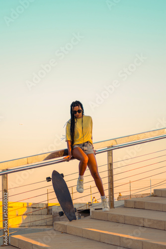 Pretty young woman with brown skin with long black braids standing with a skateboard by her side dressed in warm colored summer clothes on a concrete stairs on the seafront at summer sunset.
