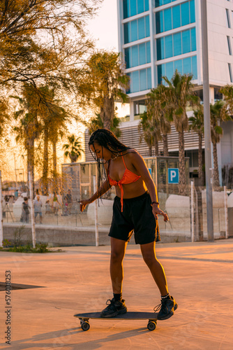Pretty young woman with brown skin with long black braids skating with her skateboard dressed in an orange bikini and black shorts having fun on the seafront in the summer sunset in the warm light.