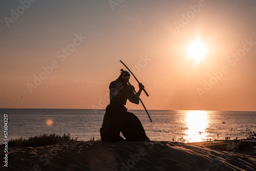 A Japanese warrior in a traditional kimano armed with a katana sword on a sandy shore next to the ocean during sunset. A man from medieval Asia. Reconstruction of cultural heritage. Culture in Japan. 