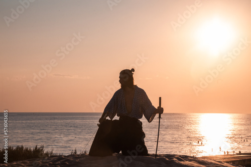 A Japanese warrior in a traditional kimano armed with a katana sword on a sandy shore next to the ocean during sunset. A man from medieval Asia. Reconstruction of cultural heritage. Culture in Japan.  photo