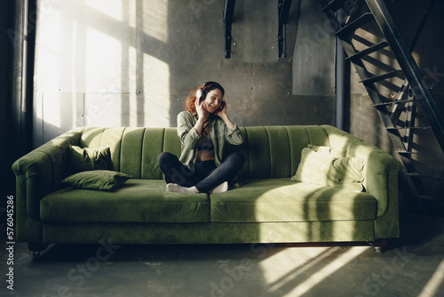 Young adult woman in headphones with closed eyes on sofa in lotus position