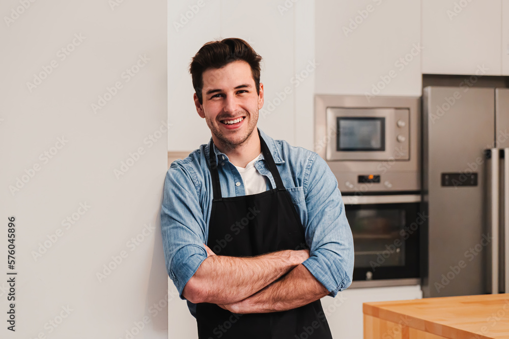 Attractive caucasian man wearing an apron standing in the kitchen, smiling and crossing arms looking at camera. Front view of happy handsome male leaning against a wall at home. High quality photo