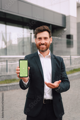 Smartphone With Blank Screen In Businessman Hand, Smiling Middle Eastern Male Entrepreneur Showing For Mobile App Or Website Advertisement While Posing Outdoors, Mockup