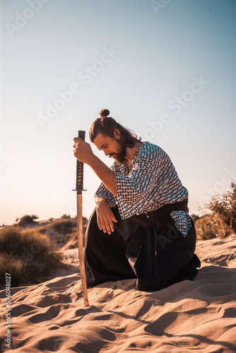 A Japanese warrior in a traditional kimano armed with a katana sword on a sandy shore next to the ocean during sunset. A man from medieval Asia. Reconstruction of cultural heritage. Culture in Japan.  photo