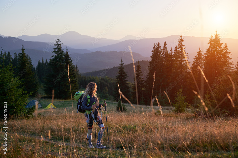 Woman traveller hiking outdoors at sunset. Sporty, slim woman traveling in mountains. Female tourist carrying backpack, using trekking sticks, admiring landscape. Concept of harmony with nature. - obrazy, fototapety, plakaty 