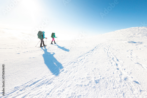 Two women on a winter hike. Girlfriends with trekking sticks go along a snow-covered mountain path. Girls with backpacks and snowshoes travel together.