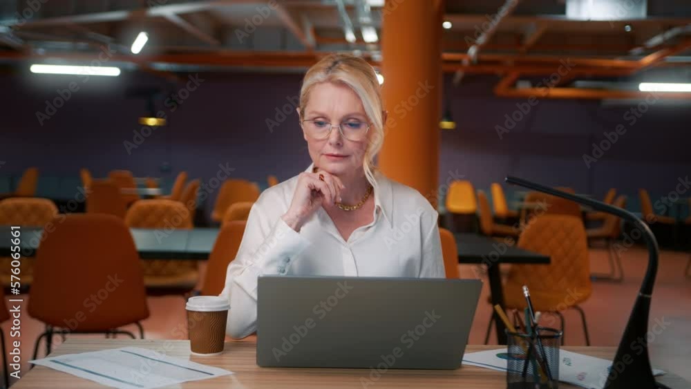 Lady boss in eye wear working on project in modern loft style empty office on motion background. Mature attractive woman in glasses working late. Business woman 50s typing reading an email on laptop Lady boss in eye wear working on project in modern loft style empty office on motion background. Mature attractive woman in glasses working late. Business woman 50s typing reading an email on laptop
