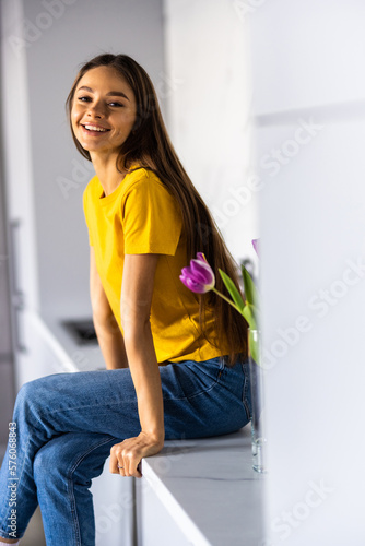 Beautiful young smiling woman working on laptop and drinking coffee while sitting on the countertop in the kitchen at home.
