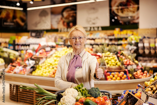 A senior woman is purchasing at the supermarket and giving thumbs up while smiling at the camera.