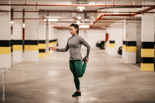 A muscular sportswoman is balancing on one foot and stretching in underground garage.