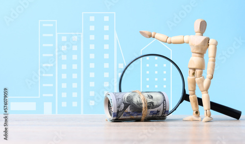 Bundle of paper dollars, magnifying glass and wooden marionette doll on blue background, concept of rising real estate prices, high mortgage rates photo